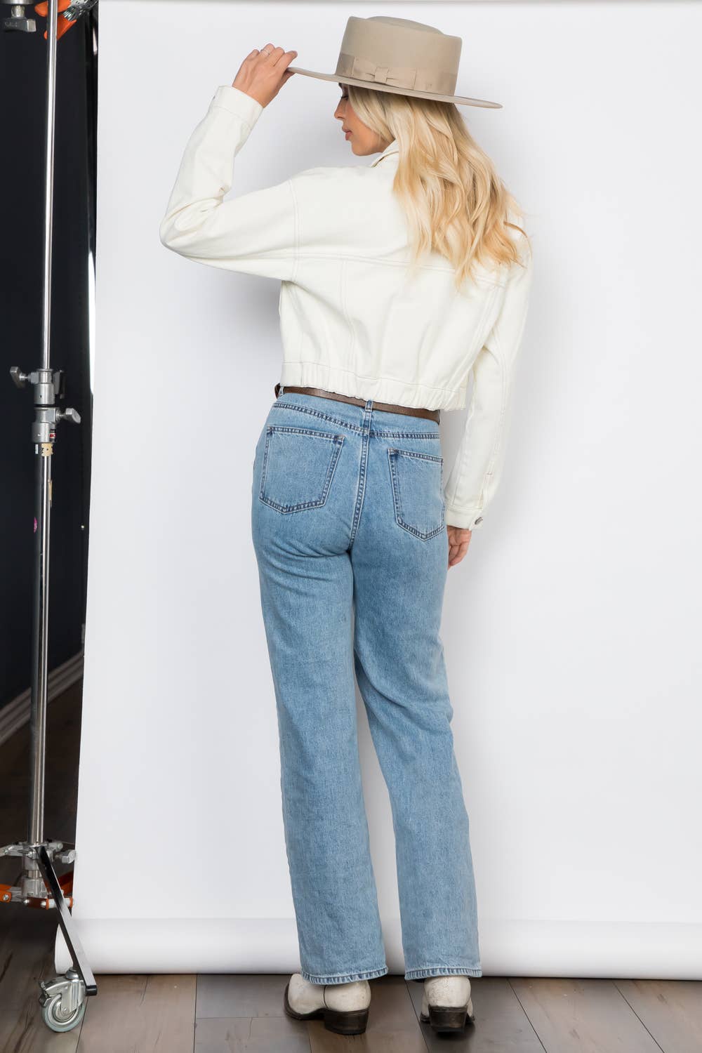 Cropped Ecru Denim Jacket Outerwear by 26 International back on model with left arm bent toward head and hand touching brim of hat, model wearing jeans and boots