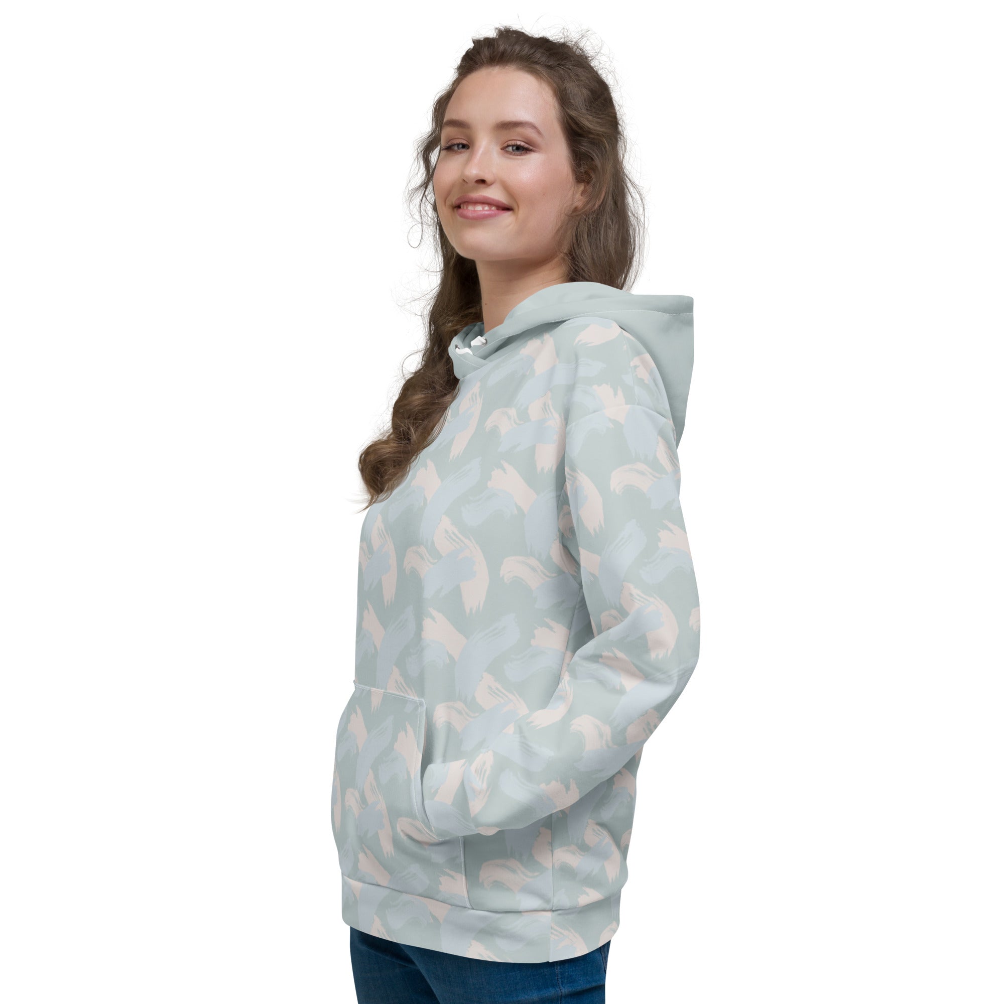 Woman wearing a light blue hoodie with abstract pattern of paint strokes in multiple colors and solid hood with pull strings tucked in facing front right and looking at camera smiling with hands in central pocket of hoodie