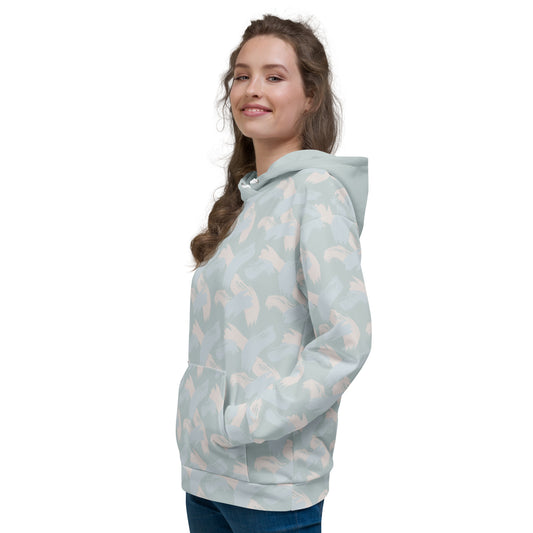Woman wearing a light blue hoodie with abstract pattern of paint strokes in multiple colors and solid hood with pull strings tucked in facing front right and looking at camera smiling with hands in central pocket of hoodie