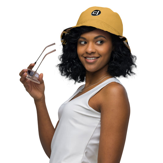 Person wearing a yellow mustard reversible interior bucket hat and holding sunglasses while looking left, logo is on interior front with alternate cheetah paint pattern showing on underside brim 