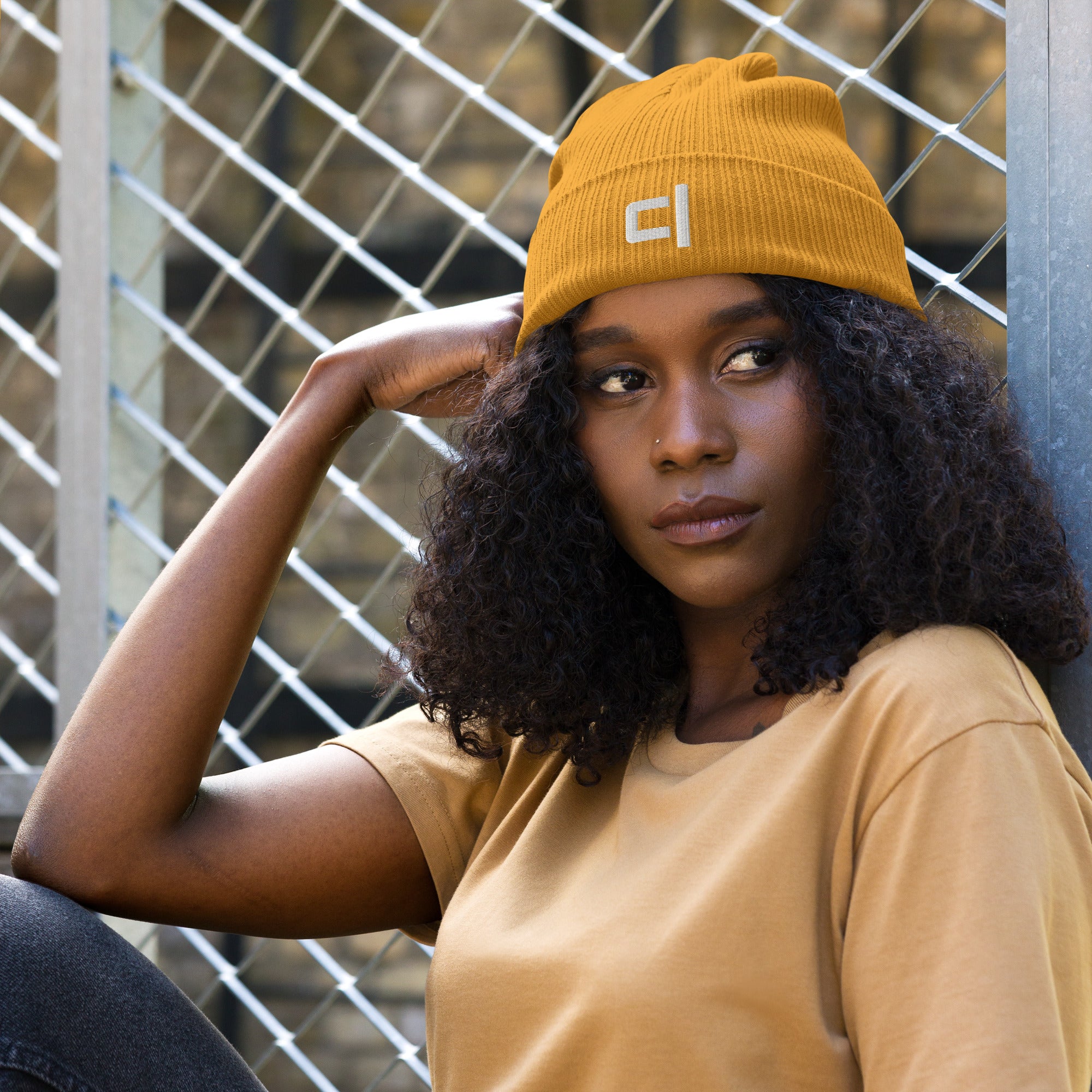 C&L Signature Organic Ribbed Beanie Beanie by Carl & Lyn mustard front view with embroidered Carl & Lyn logo in white and person sitting with head leaning toward right arm looking to left in front of cyclone fence